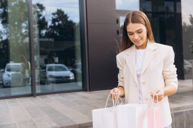 Modern bir mağazanın önünde yürürken elinde alışveriş torbalarıyla gülümseyen bir kadın. Şehirdeki eğlence ve moda keşifleriyle dolu keyifli bir şehir alışveriş deneyimi..
