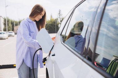 Genç bir kadın, çevre dostu yaşam tarzı ve sürdürülebilirliği yansıtan, bir kamu istasyonunda elektrikli bir araç şarj ediyor. Güneşli açık hava manzarası sahneye olumlu, fütüristik bir hava katıyor..