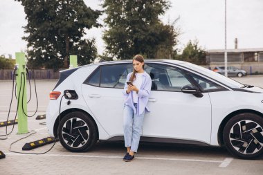 Genç bir kadın akıllı telefonunu kullanırken, şehir ortamında çevre dostu seyahat ve sürdürülebilir enerjiyi kucaklarken, bir şehir istasyonunda elektrikli arabasını şarj ediyor..