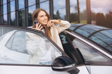 Kendine güvenen genç bir kadın arabasının yanında duruyor, telefon konuşmasının keyfini çıkarıyor. Rahat ve mutlu görünüyor, modern bir şehir geçmişine karşı gelişigüzel giyinmiş..