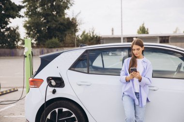 Genç bir kadın şarj istasyonunda elektrikli arabasına yaslanır, akıllı telefonuyla meşgul olur. Sahnede modern teknoloji, sürdürülebilirlik ve elektrikli araçların kolaylığı vurgulanıyor.