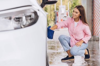 Smiling young woman cleaning her car with a blue cloth at a self-service car wash. She's dressed casually in a pink shirt and jeans, enjoying the sunny day while maintaining her vehicle. clipart