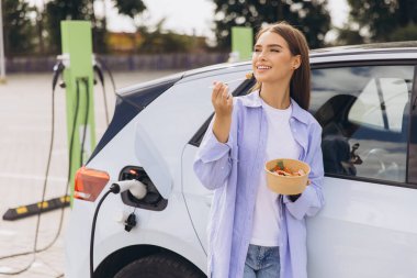 Genç bir kadın açık hava istasyonunda elektrikli arabası şarj edilirken sağlıklı bir salatanın tadını çıkarıyor. Sahne, sürdürülebilirliği, sağlıklı yaşam tarzını ve modern teknolojiyi huzur ortamında yansıtıyor..