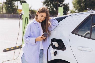 Genç bir kadın şarj istasyonunda akıllı telefonunu kullanarak elektrikli arabasına yaslanıyor. Kurulumda modern teknoloji, sürdürülebilirlik ve kolaylık vurgulanıyor.