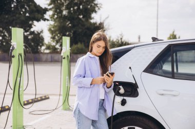 Genç bir kadın şarj istasyonunda akıllı telefonunu kullanarak elektrikli arabasına yaslanıyor. Kurulumda modern teknoloji, sürdürülebilirlik ve kolaylık vurgulanıyor.