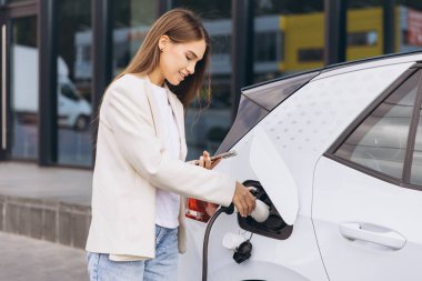 Açık hava şarj istasyonunda elektrikli arabasını şarj ederken akıllı telefon kullanan genç bir kadın. Teknolojiyi konfor ve çevre dostu ulaşım için kullanıyor gibi görünüyor..