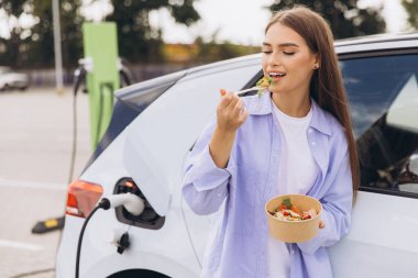 Çağdaş şarj istasyonunda elektrikli arabası şarj olurken genç bir kadın taze salatanın tadını çıkarıyor. Bu resim modern yaşam tarzını ve sürdürülebilir yaşamı yakalar..
