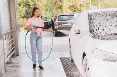 Genç bir kadın bir self-servis istasyonunda arabasını güvenli bir şekilde yıkıyor. Doğa ve diğer arabalarla çevrili bir otoparkta sabun sıkmak için hortum kullanıyor..