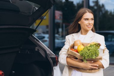 Genç bir kadın, süpermarketin otoparkındaki bagajına içi taze yiyecek dolu kağıt torbaları doldururken gülümsüyor..