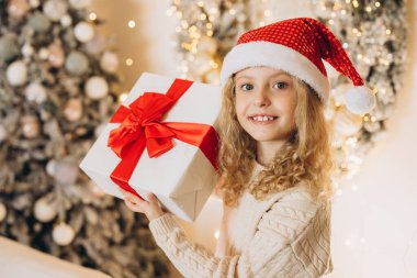 Noel Baba şapkası takan neşeli genç bir kız güzel bir Noel hediyesi tutuyor. Büyük kırmızı bir fiyonk, bayram süslemeleri ve parlak ışıklarla çevrili..