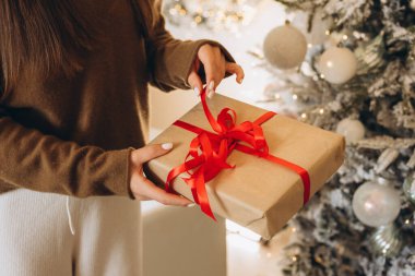 Samimi bir süveter giyen bir kadın, kırmızı kurdeleyle süslenmiş bir hediye kutusu tutuyor. Güzel dekore edilmiş bir Noel ağacının yanında duruyor. Sahne, bayram ruhunu ve hediye vermenin neşesini yakalıyor..