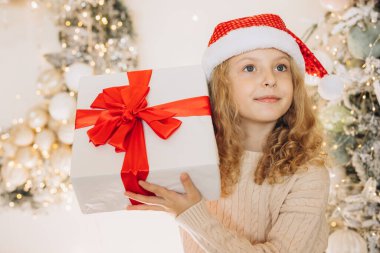 Noel Baba şapkalı neşeli genç bir kız güzel bir Noel hediyesi tutuyor kırmızı kurdeleli, etrafı şenlikli süslemeler ve ışıl ışıl ağaçlarla çevrili..