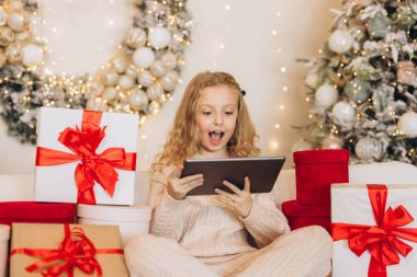 Neşeli küçük bir kız Noel hediyeleriyle çevrili bir kanepede oturuyor ve mutlu bir şekilde tablet kullanıyor. Şenlikli dekorasyonlar sıcak ve neşeli bir tatil ortamı yaratıyor..