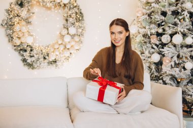 A woman with a smile opens a beautifully wrapped gift in a cozy Christmas setting, surrounded by festive decorations. The warm atmosphere captures the joy and excitement of holiday celebrations. clipart