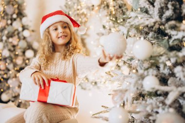 Noel Baba şapkası takan neşeli bir çocuk güzel süslenmiş bir Noel ağacının yanında bir hediye tutuyor. Bayram sezonunun coşkusunu ve coşkusunu simgeliyor..