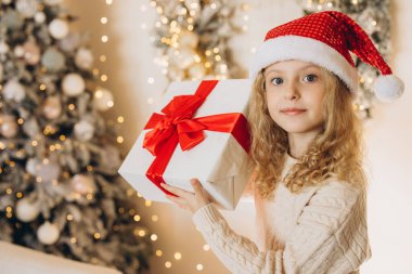 Noel Baba şapkası takan neşeli genç bir kız güzel bir Noel hediyesi tutuyor. Büyük kırmızı bir fiyonk, bayram süslemeleri ve parlak ışıklarla çevrili..