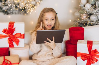 Neşeli bir çocuk güzel paketlenmiş Noel hediyelerinin arasında heyecanlı bir şekilde tablet kullanıyor. Şenlik atmosferi parlak ışıklar ve arka plandaki tatil dekorasyonlarıyla güçlendiriliyor..