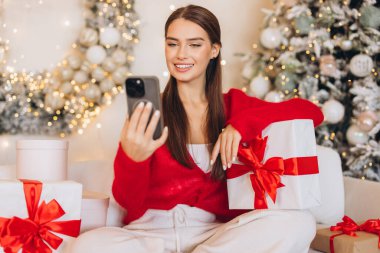 A joyful woman sits by a decorated Christmas tree, holding a gift and texting on her phone, surrounded by festive lights and wrapped presents.