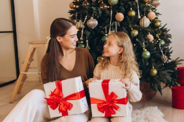 Neşeli bir anne ve kız, süslenmiş bir Noel ağacının yanında oturur, güzel hediyeler alır, neşeli gülümsemeler ve bayram heyecanı paylaşırlar..