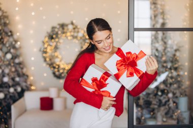 Kırmızı kazaklı, gülümseyen bir kadın, Noel hediyelerini güzel dekore edilmiş bir odada, neşeli bir atmosferle kucaklıyor ve Noel ruhunu sergiliyor..