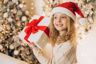 Noel Baba şapkası takan neşeli genç bir kız güzel bir Noel hediyesi tutuyor. Büyük kırmızı bir fiyonk, bayram süslemeleri ve parlak ışıklarla çevrili..
