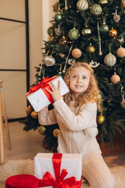 Kıvırcık saçlı neşeli bir çocuk güzel dekore edilmiş bir Noel ağacının önünde kırmızı ve beyaz bir hediye açar. Sıcaklık, neşe ve şenlik havası yayar..