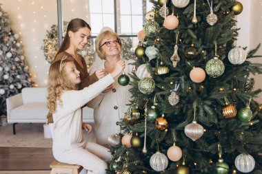 Aralarında büyükanne, anne ve genç bir kızın da bulunduğu üç nesildir sıcak aydınlatılmış oturma odasındaki süslemelerle bir Noel ağacını neşeyle süslediler..