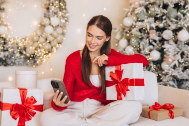 A joyful woman in a cozy red sweater enjoys her Christmas gifts while using her smartphone beside a beautifully decorated tree. Festive atmosphere with holiday lights and wrapped presents.