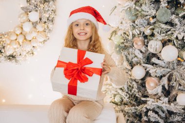Noel Baba şapkası takan neşeli bir kız güzel dekore edilmiş bir Noel ağacının önünde oturuyor, elinde büyük kırmızı bir fiyonk ile bir hediye tutuyor ve gülümsüyor..