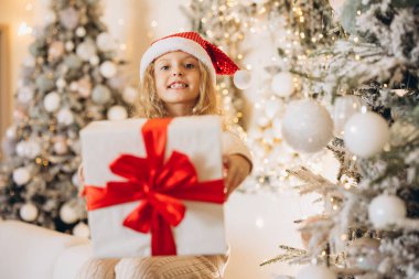 Noel Baba şapkalı mutlu bir çocuk, süslerle süslenmiş parlak bir Noel ağacının yanında kırmızı kurdeleli güzel bir hediye tutar. Sahne neşeli sevinç ve tatil sıcaklığını yakalıyor..