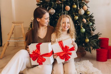 Neşeli bir anne ve kız, süslenmiş bir Noel ağacının yanında oturur, güzel hediyeler alır, neşeli gülümsemeler ve bayram heyecanı paylaşırlar..