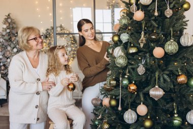 Aralarında büyükanne, anne ve genç bir kızın da bulunduğu üç nesildir sıcak aydınlatılmış oturma odasındaki süslemelerle bir Noel ağacını neşeyle süslediler..