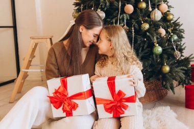 Bir anne ve kızı, güzel süslenmiş bir Noel ağacının önünde hediyelerini verirken sevinçle kucaklaşırlar. Şenlikli atmosfer, tatil sezonunun sıcaklığını ve sevgisini vurguluyor..