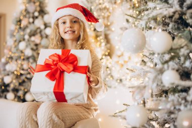 Noel Baba şapkalı mutlu bir çocuk, süslerle süslenmiş parlak bir Noel ağacının yanında kırmızı kurdeleli güzel bir hediye tutar. Sahne neşeli sevinç ve tatil sıcaklığını yakalıyor..