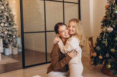 Neşeli bir anne ve kız, güzel dekore edilmiş bir Noel ağacının yanında sıcak bir kucaklaşmayı paylaşır. Tatil sezonunda sıcacık bir evde sevgi ve birlikteliği sembolize eder..