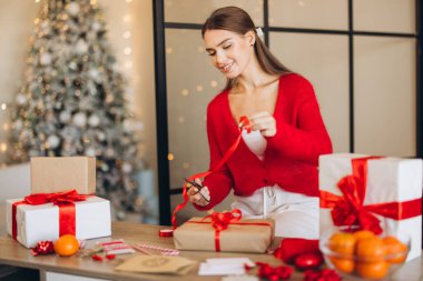 Kırmızı kazaklı bir kadın, süslü bir Noel ağacının ve parıldayan ışıkların yer aldığı, neşe ve tatil ruhunu yansıtan kırmızı kurdeleli bir odada hediyeler sunuyor..