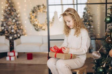 Genç bir kız, Noel hediyesini güzel dekore edilmiş bir oturma odasında, Noel ışıkları ve süslemelerle çevrili bir şekilde açar. Sahne, festival sezonunun sıcaklığını ve neşesini yakalar..