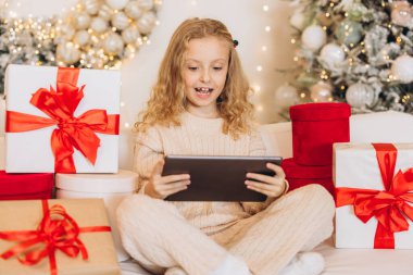 Neşeli küçük bir kız Noel hediyeleriyle çevrili bir kanepede oturuyor ve mutlu bir şekilde tablet kullanıyor. Şenlikli dekorasyonlar sıcak ve neşeli bir tatil ortamı yaratıyor..