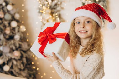 Noel Baba şapkası takan neşeli genç bir kız güzel bir Noel hediyesi tutuyor. Büyük kırmızı bir fiyonk, bayram süslemeleri ve parlak ışıklarla çevrili..