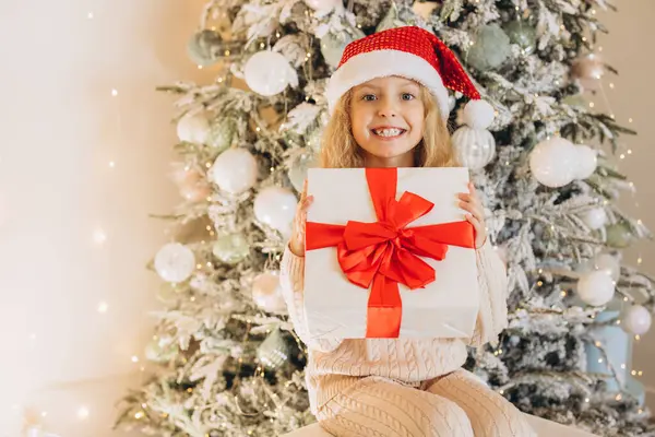 Noel Baba şapkası takan neşeli bir kız güzel dekore edilmiş bir Noel ağacının önünde oturuyor, elinde büyük kırmızı bir fiyonk ile bir hediye tutuyor ve gülümsüyor..