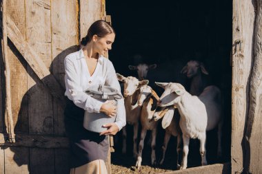 A young woman stands in a barn door holding a metal milk can, surrounded by goats. The warm sunlight creates a serene farm atmosphere. clipart