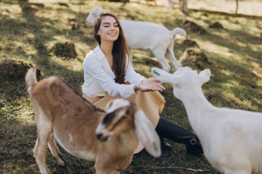Neşeli genç bir kadın, güneşli bir günde keçilerle iletişim kurarken gülümser, doğanın ve hayvan arkadaşlığının neşesini gösterir..