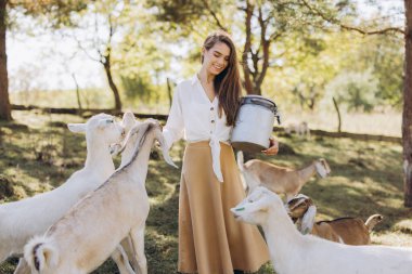 Elinde kova tutan neşeli genç bir kadın huzurlu bir çiftlik ortamında keçileri besliyor. Bereketli yeşillik ve güneş ışığı kırsal yaşamı ve doğayla bağlantıyı gösteren barışçıl bir atmosfer yaratır..