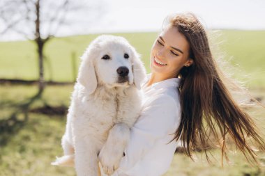 Neşeli genç bir kadın, güneşli yeşil bir alanda tüylü beyaz köpeğini kucaklıyor. İfadeleri mutluluğu ve arkadaşlığı yansıtıyor, sakin bir açık hava keyfi anı sergiliyor..