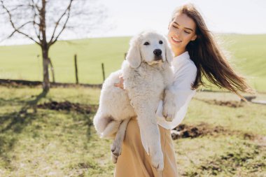 Neşeli genç bir kadın, güneşli bir alanda tüylü beyaz bir köpek yavrusunu sevgiyle kucağına alır. Açık havada huzurlu bir ortamda neşe ve arkadaşlığı somutlaştırır..