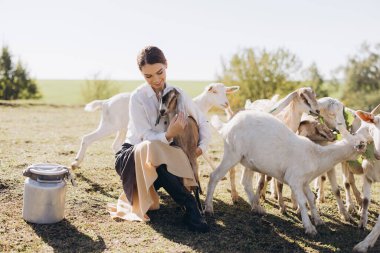 Bir kadın, güneşli bir kırsal alanda keçilerin arasında oturmuş, küçük bir keçiyi kucaklıyor. Sahne sükunet, doğa ile bağlantı ve pastoral cazibe yayar..