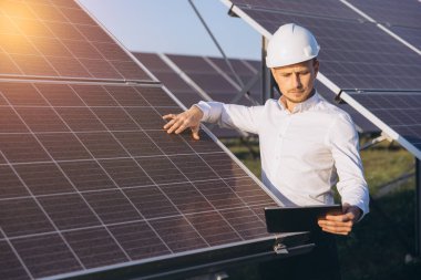 Beyaz gömlekli ve kasklı profesyonel mühendis güneş panellerini inceliyor. Güneş ışığı, yenilenebilir enerji teknolojisi ve sürdürülebilir çözümlere odaklanılmasını vurguluyor. Temiz enerjide yenilik.