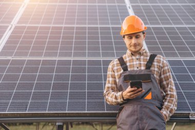 Ekose gömlekli ve şapkalı bir erkek mühendis güneş panellerine yaslanırken akıllı telefon kullanıyor. Görüntü, teknoloji, yenilenebilir enerji ve sürdürülebilir endüstri uygulamaları kavramlarını iletir.