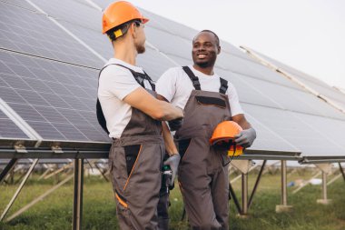 Koruyucu giysili iki mühendis güneş çiftliğinde çalışmayı tartışıyor. Güneş panellerini denetliyor, takım çalışmasını ve sürdürülebilir enerji gelişimini sembolize ediyorlar..