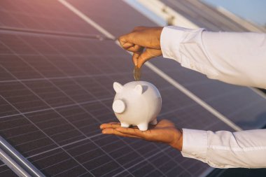 Güneş panellerinin önündeki kumbara içine bozuk para koyan biri yenilenebilir enerji tasarrufunu simgeliyor. Finansal büyüme ve sürdürülebilirlik kavramları vurgulandı.